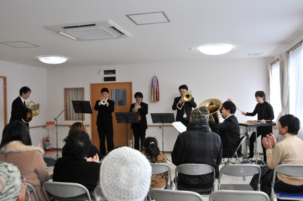 仮設・地域の集会のような広間で、前方の聴衆に向けて金管グループが演奏している様子